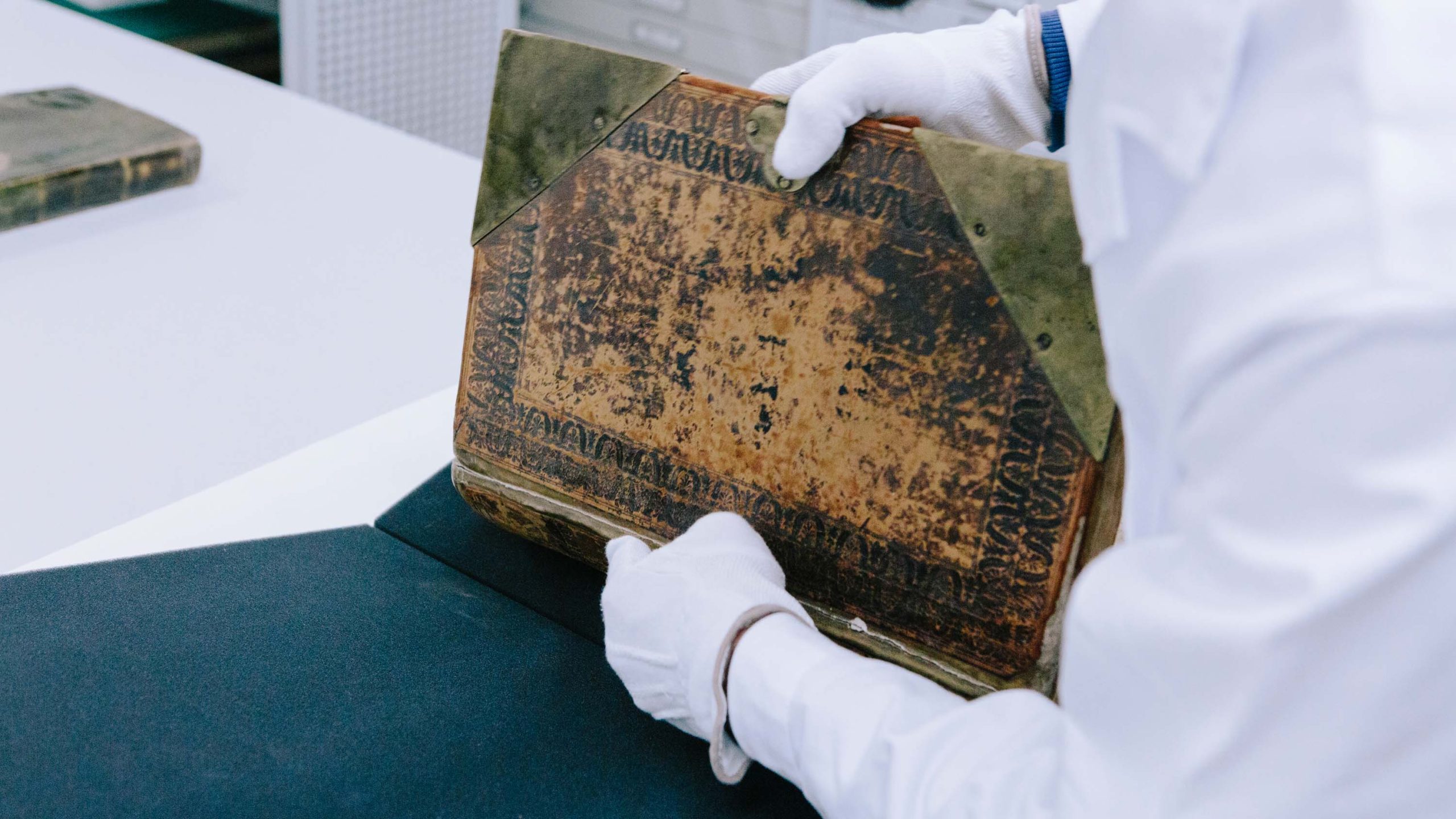 Eine Person in einem weißen Arbeitsmantel und mit weißen Handschuhen packt auf einem Tisch ein historisches Buch aus einer Aufbewahrungsbox. Der Bucheinband sieht sehr als aus, der Buchrücken ist etwas veschädigt.