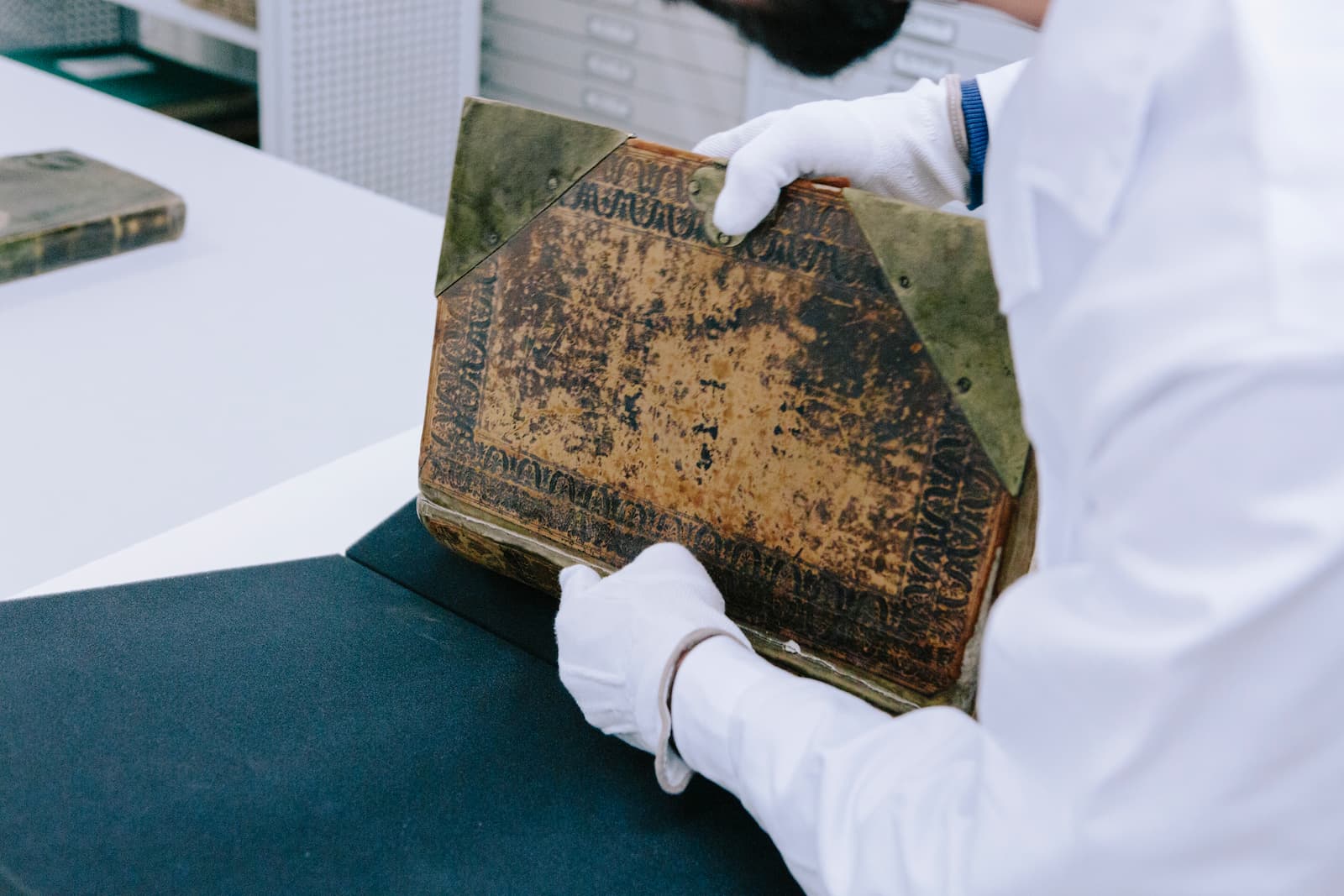 Eine Person in einem weißen Arbeitsmantel und mit weißen Handschuhen packt auf einem Tisch ein historisches Buch aus einer Aufbewahrungsbox. Der Bucheinband sieht sehr als aus, der Buchrücken ist etwas veschädigt.