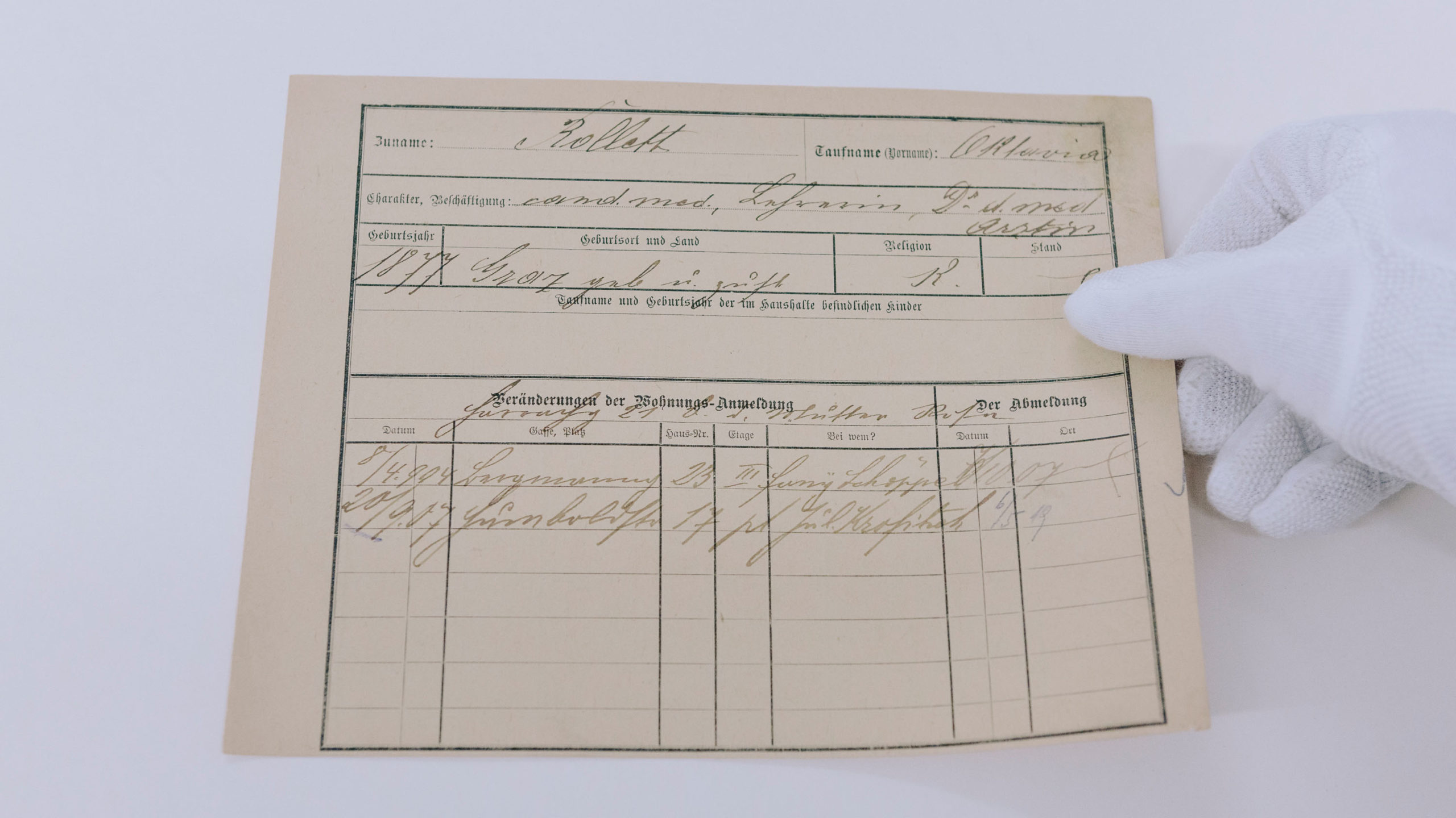 Eine Hand mit weißen Stoffhandschuhen hält einen alten Meldezettel von Oktavia Rollett, auf dem handschriftlichen Daten in Kurrent vermerkt sind.