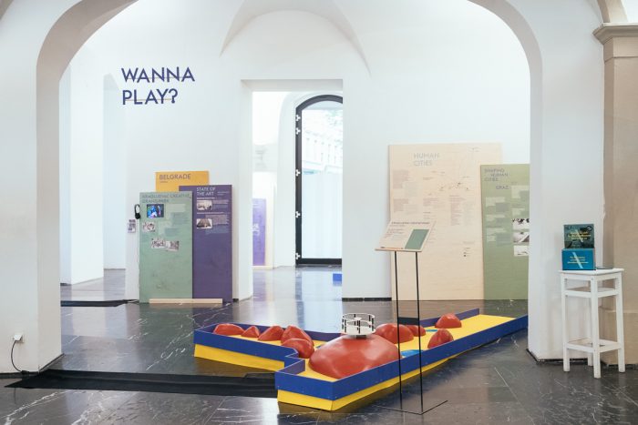 Ausstellungsraum mit Tafeln von Shaping Human Cities, bunter Spielstruktur und WANNA PLAY? an der Wand.