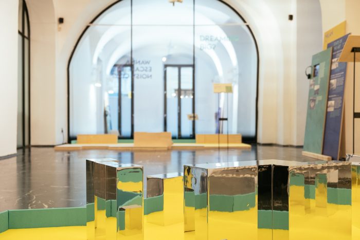 Reflektierende Metallblöcke auf einer gelben Plattform erforschen das städtische Design in einem modernen Ausstellungsraum.