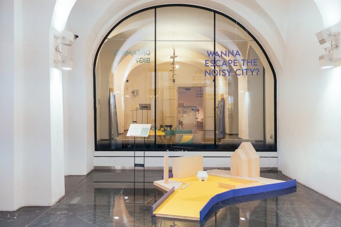 Modernes Kunstexponat mit Stadtgestaltungsmodellen, Glaswand und Text: WILLST DU DER LAUTEN STADT ENTKOMMEN?.