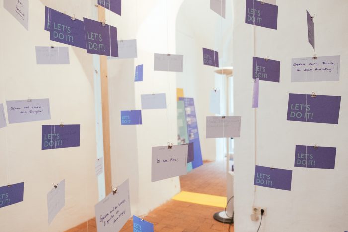 Karten mit handschriftlichen Notizen und "Lets Do It!"-Schildern unterstreichen das urbane Design in einem Raum mit weißen Wänden.