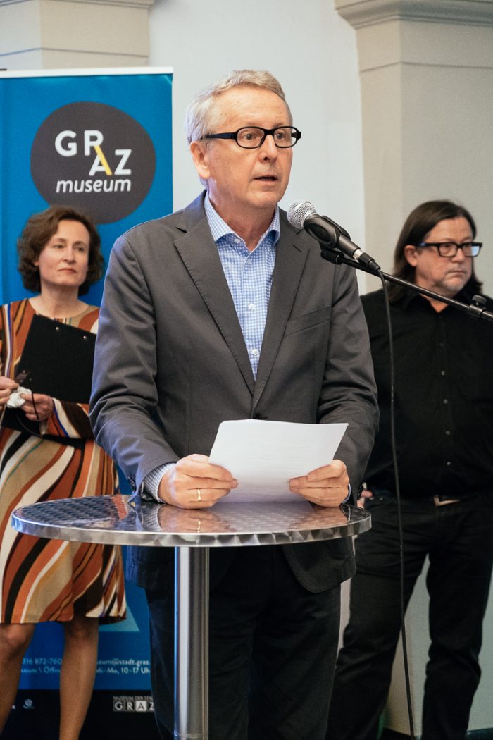 Ein Mann im Anzug spricht auf dem Podium über Stadtplanung, zwei Personen stehen hinter ihm bei einer Veranstaltung in einer Halle.