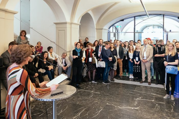 Eine Frau spricht auf einem Podium zu einer Menschenmenge in einer Lobby, die unter gewölbten Decken menschliche Städte formt.