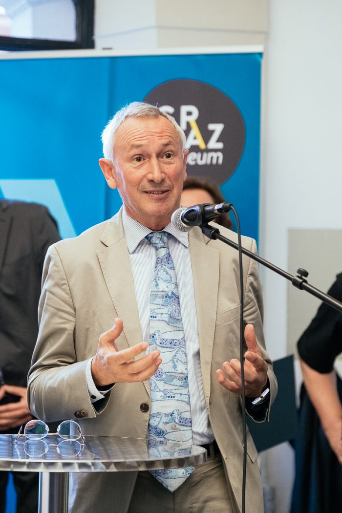 Ein Mann in einem hellbraunen Anzug diskutiert auf einem Podium mit Mikrofon während einer Veranstaltung in einer Halle über Stadtplanung.
