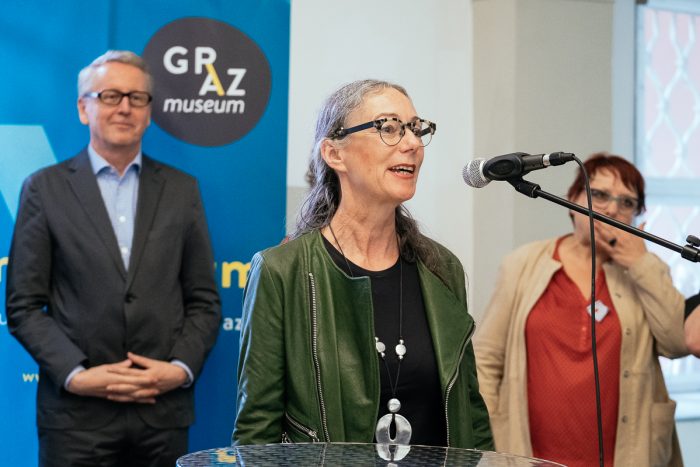 Frau, die an einem Mikrofon über Stadtplanung spricht, mit zwei Personen hinter ihr bei einer Veranstaltung in einer Halle.