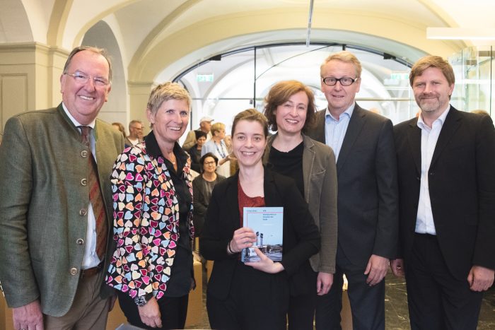 Sechs Erwachsene stehen in einem Raum, eine Frau in der Mitte hält ein Buch und lächelt in die Kamera.