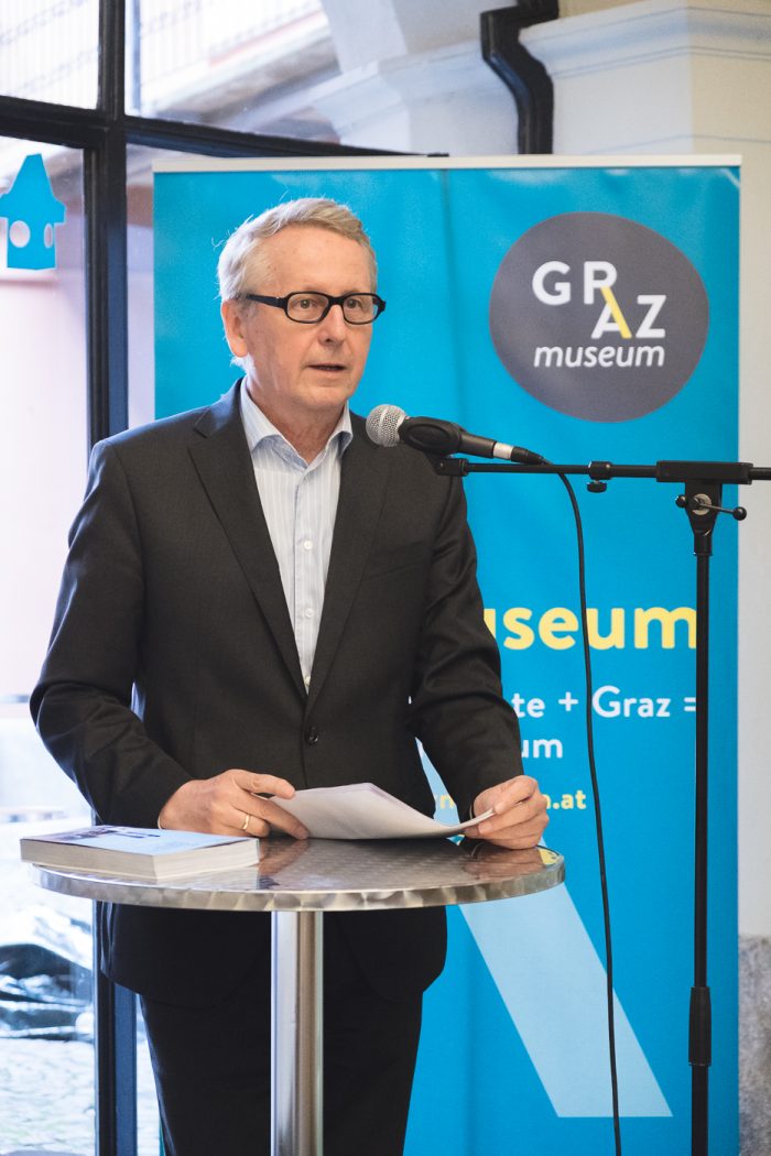 Ein Mann im Anzug spricht an einem Podium mit einem Mikrofon vor einem Schild des Graz Museums.