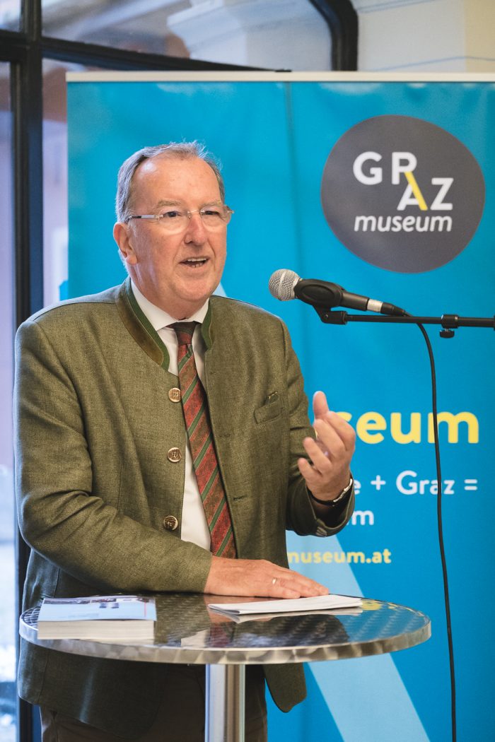 Ein älterer Mann spricht auf einem Podium vor einem Transparent des Graz Museums.
