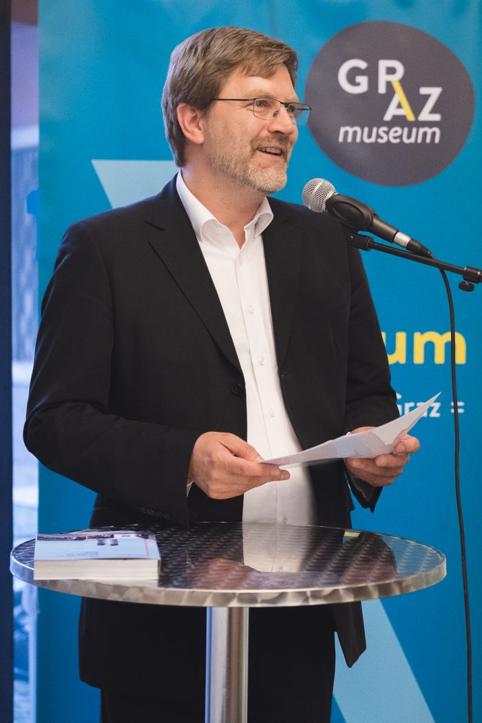Ein Mann im Anzug spricht an einem Podium mit einem Mikrofon bei einer Veranstaltung des Graz Museums.