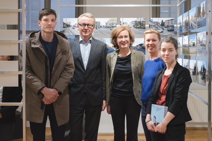 Fünf Personen stehen in einem Innenraum vor einer Wand mit Fotos und posieren für ein Gruppenfoto.