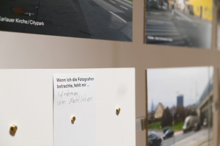 Eine Notiz in deutscher Sprache ist unter die ausgestellten Stadtansichten in einer Ausstellung gepinnt.