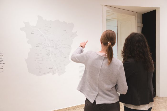 Zwei Personen betrachten und diskutieren eine Karte, die an einer weißen Wand in einem Galerieraum angebracht ist.