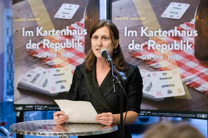 Eine Frau spricht an einem Podium in Graz mit Plakaten mit der Aufschrift Im Kartenhaus der Republik 1918-1938.