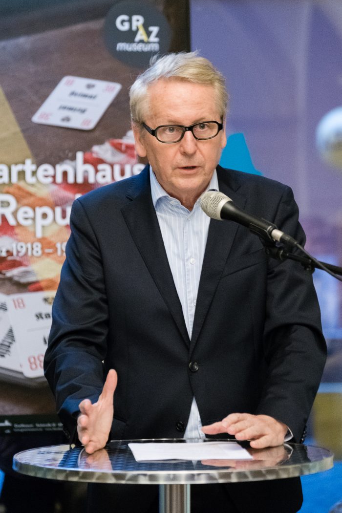 Ein Mann im Anzug spricht an einem Podium, im Hintergrund ein Plakat mit dem Hinweis auf die Republik Graz 1918-1938.