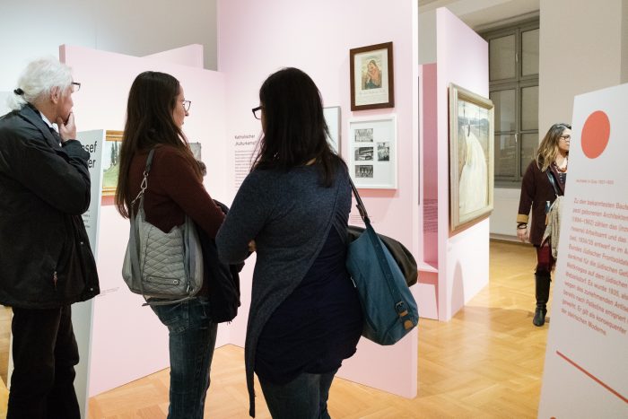 Vier Personen betrachten in einer hell erleuchteten Grazer Galerie mit rosa Wänden Kunst aus der Zeit der Republik von 1918-1938.