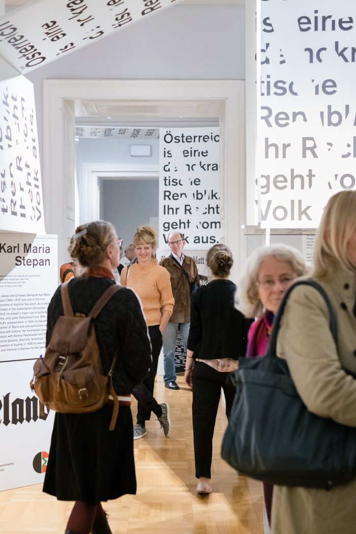 Menschen erkunden eine Ausstellung der Republik 1918-1938 in Graz, umgeben von mit Text bedeckten Wänden.