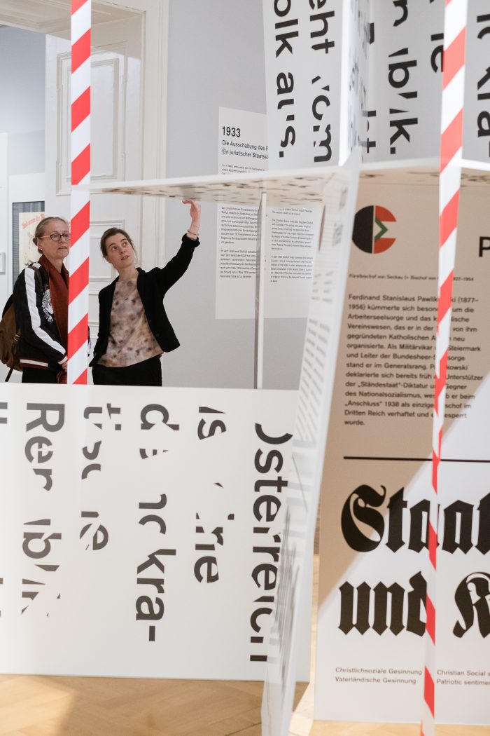 Zwei Personen betrachten das Grazer Kartenhaus der Republik, 1918-1938, mit Texttafeln und rot gestreiften Masten.