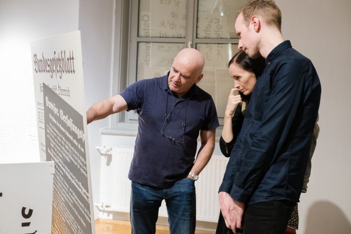 Drei Personen diskutieren über einen Text auf einer Schautafel über die Republik Graz von 1918-1938 in einer Galerie.