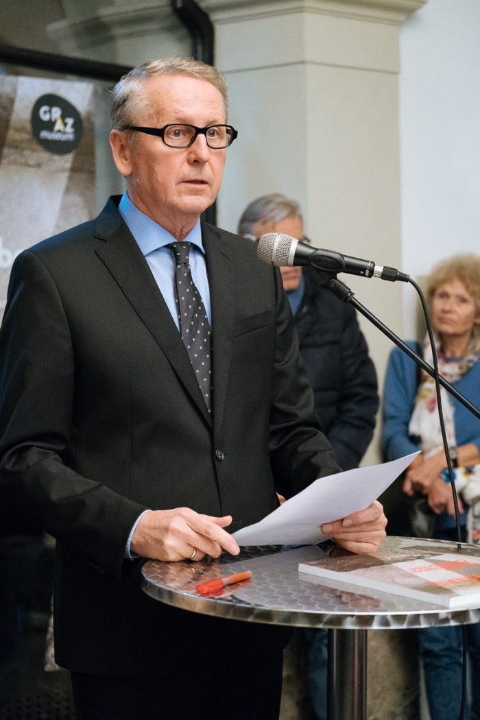 Mann im Anzug spricht am Podium über das Lager Liebenau, im Hintergrund sitzen Menschen.