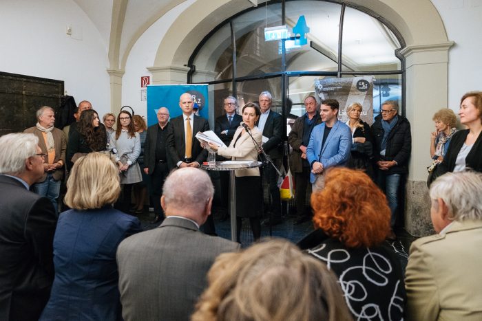Eine Frau spricht an einem Podium über das Lager Liebenau zu einem versammelten Publikum in einem formellen Innenbereich.