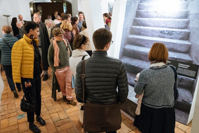Eine Gruppe bewundert eine Ausstellung über das Lager Liebenau, auf der ein großes Foto von Steinstufen zu sehen ist.