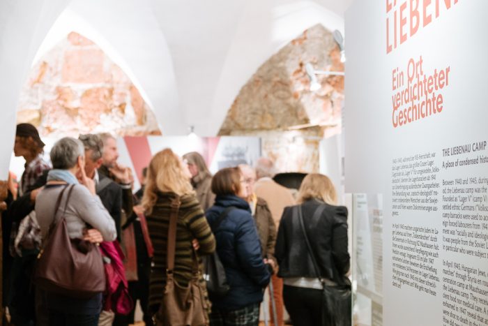 Eine Gruppe von Menschen betrachtet eine Lager Liebenau-Ausstellung mit Informationstext an einer Wand.