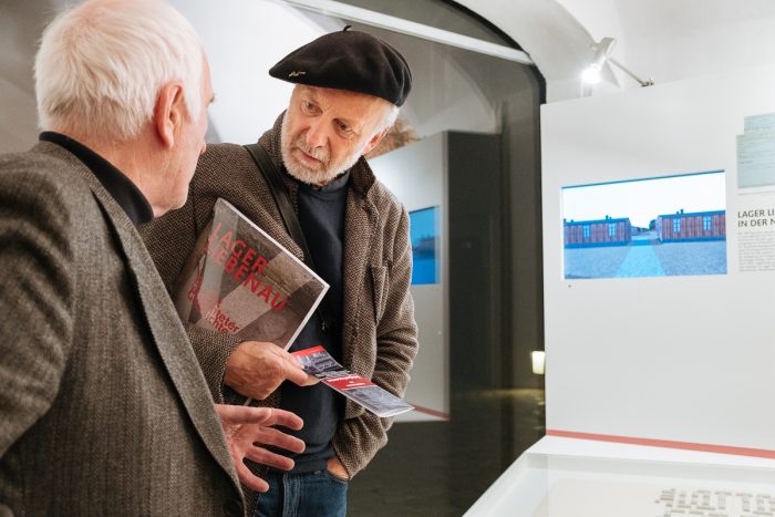 Zwei ältere Männer unterhalten sich in einem Museum, der eine hält ein Buch mit dem Titel Lager Ebensee und Lager Liebenau.