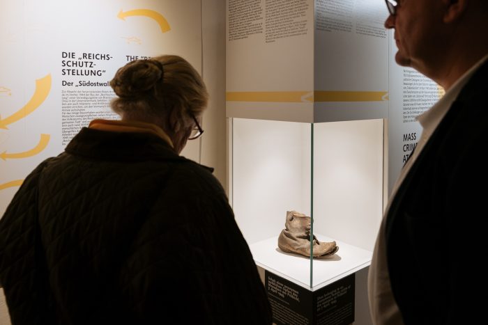 Zwei Personen betrachten einen alten Schuh aus dem Lager Liebenau in einer Glasvitrine in einer Museumsausstellung.