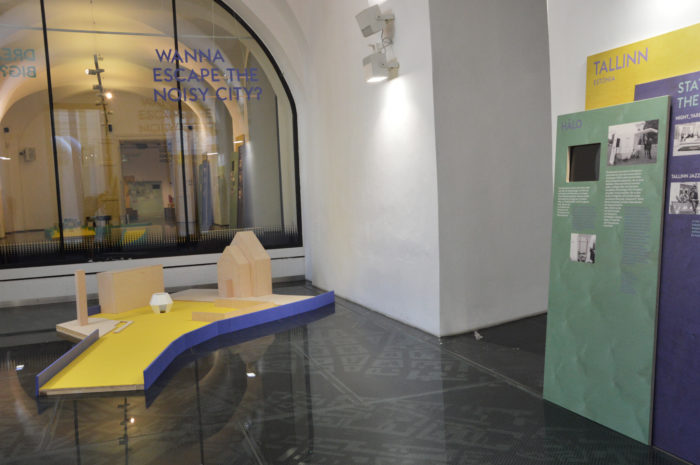 Ausstellungsdisplay mit einem Modell, einer grünen Infotafel über Tallinn und einem großen Fenster mit Text.