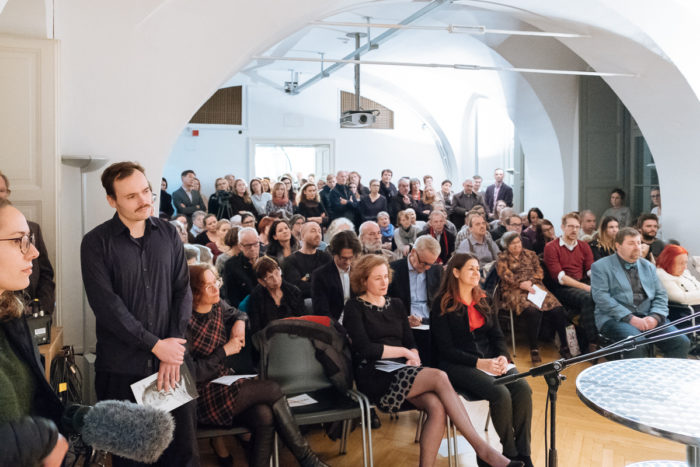 Eine große Gruppe von Menschen sitzt und steht in einem hellen Raum und nimmt an einer formellen Veranstaltung teil.