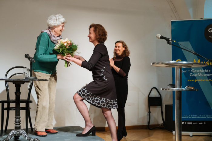 Eine Frau überreicht einer anderen Frau auf der Bühne einen Blumenstrauß, während eine dritte Frau in der Nähe applaudiert.