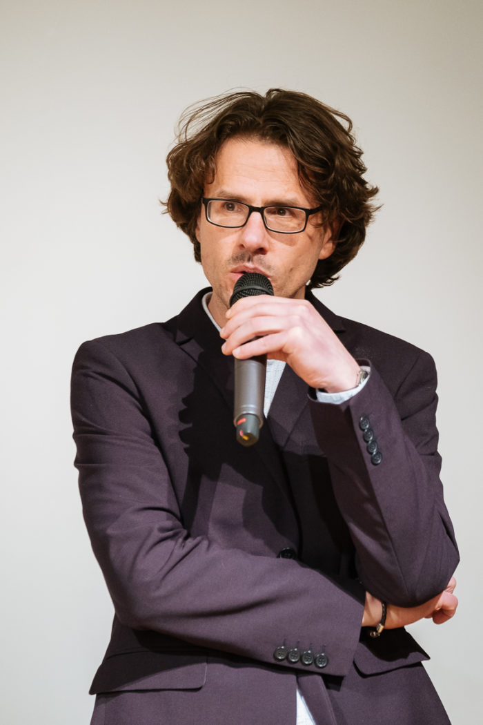 Ein Mann mit Brille und Anzugjacke spricht in ein Mikrofon vor einem schlichten Hintergrund.