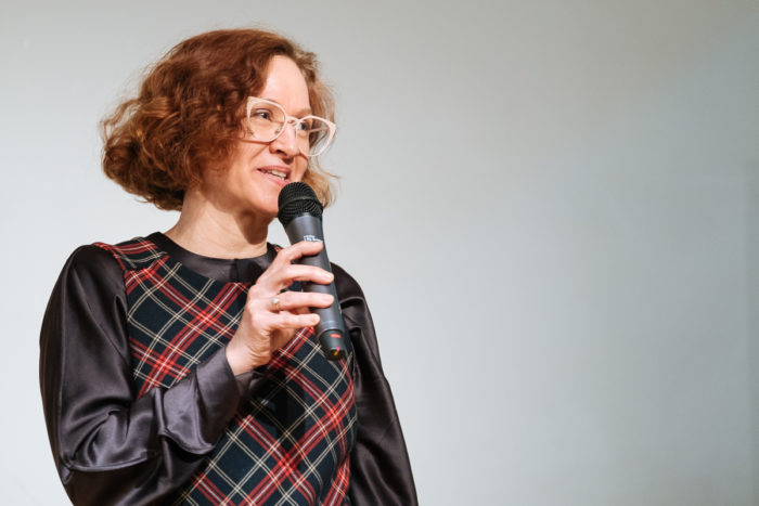 Frau mit lockigem Haar und Brille spricht in ein Mikrofon vor einem schlichten Hintergrund.