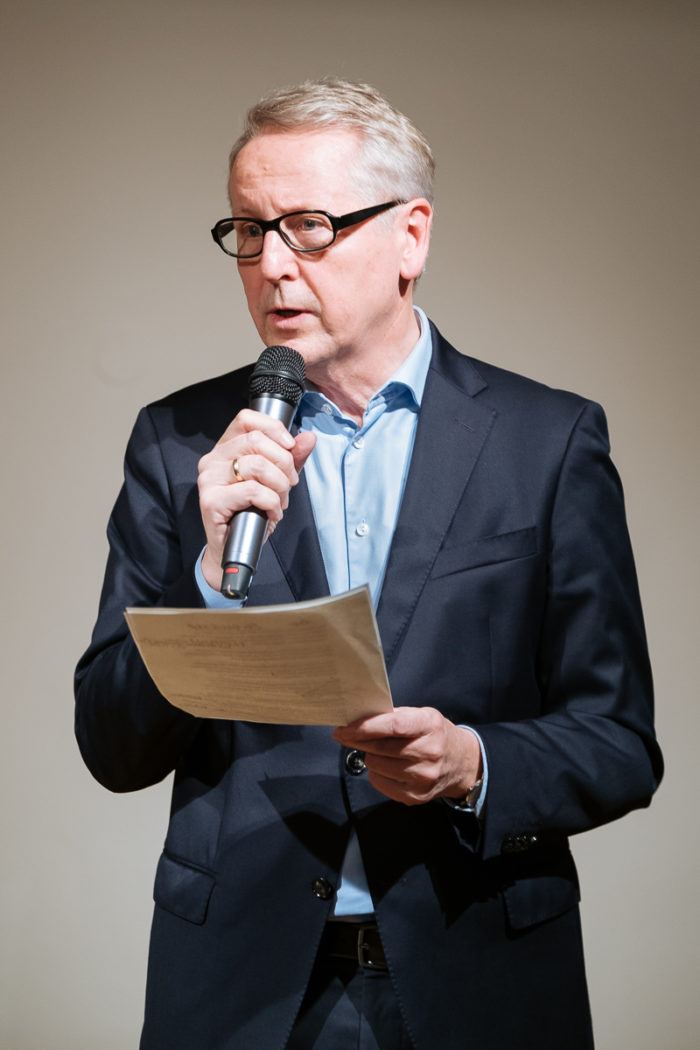 Ein Mann im Anzug und mit Brille spricht in ein Mikrofon und hält Papiere in der Hand.