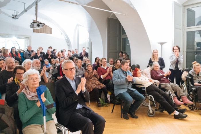 Eine große Gruppe von Menschen, darunter auch Senioren, sitzt und applaudiert bei einer Indoor-Veranstaltung.