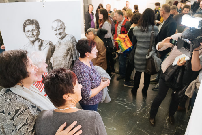 Eine Gruppe posiert für ein Foto bei einer Kunstausstellung mit einer Menschenmenge und einem großen Porträt im Hintergrund.