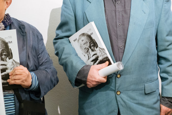 Zwei Personen in blauen Jacken halten Bücher mit einem schwarz-weißen Gesicht auf dem Einband.