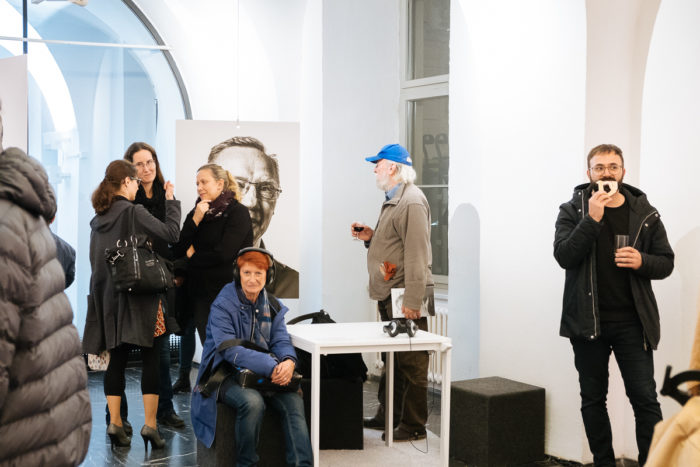 Stehende und sitzende Menschen in einer Kunstgalerie, in der Schwarz-Weiß-Porträts ausgestellt sind.