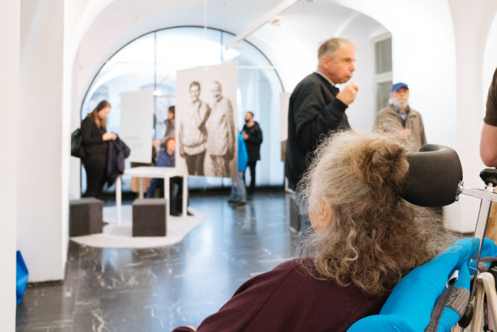 Menschen betrachten Kunst und interagieren in einer Galerie; im Vordergrund ist eine Frau im Rollstuhl zu sehen.