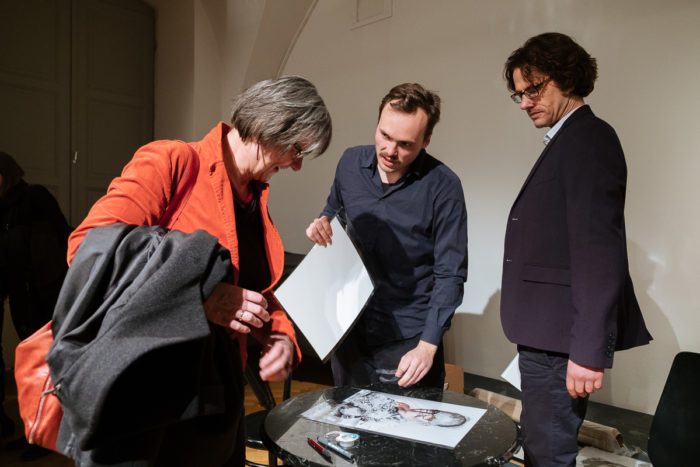 Drei Personen betrachten ein gedrucktes Foto auf einem runden Tisch in einem gut beleuchteten Raum.