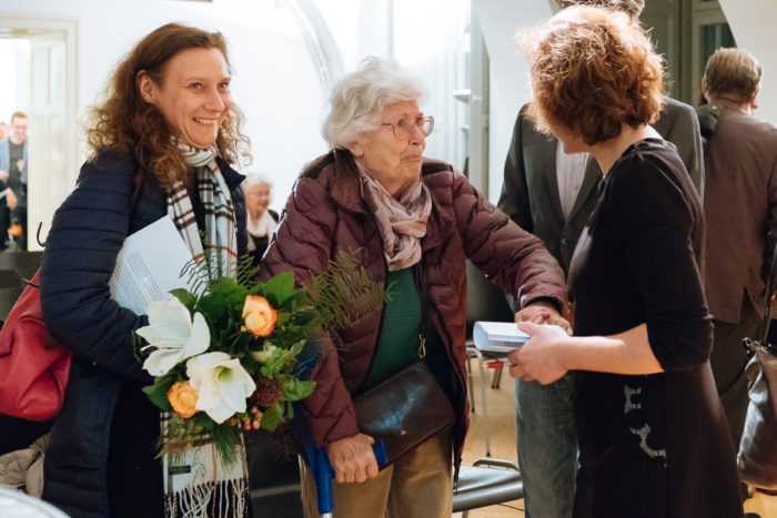 Drei Frauen stehen in einem Haus zusammen, eine hält Blumen, eine andere benutzt einen Stock und unterhält sich.