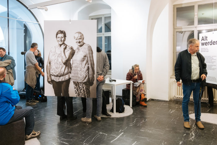 Zwei Personen halten ein großes Foto von zwei Frauen in einem Galerieraum mit anderen Personen im Hintergrund.