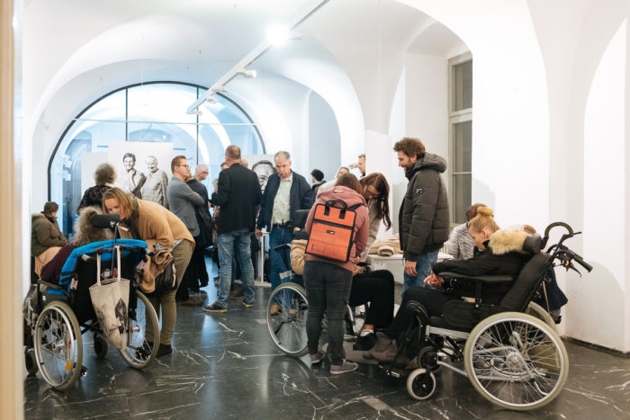 In einem hellen, gewölbten Galerieraum besuchen Menschen, darunter einige im Rollstuhl, eine Kunstausstellung.