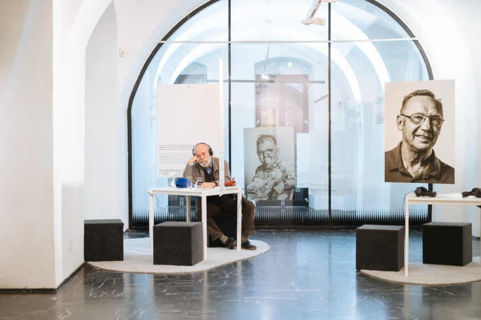 Ein Mann mit Kopfhörern sitzt an einem Tisch in einer modernen Galerie, in der Schwarz-Weiß-Porträts ausgestellt sind.