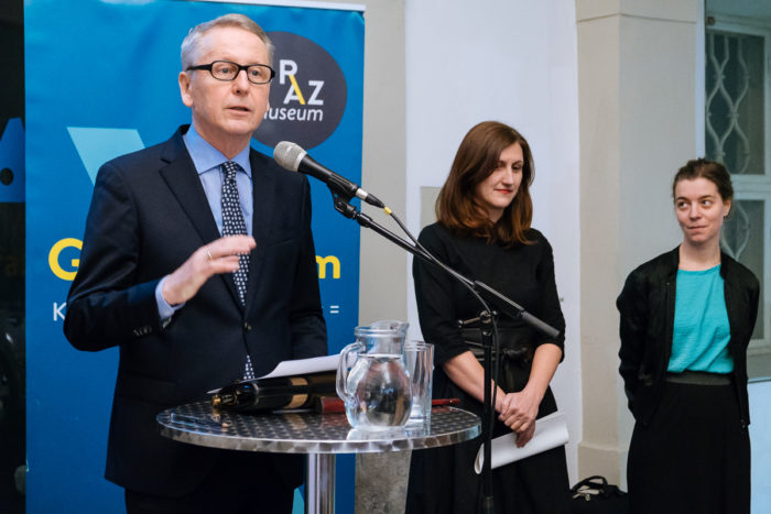 Ein Mann spricht auf einem Podium, während zwei Frauen bei einer ganz normalen anderen Indoor-Veranstaltung daneben stehen.