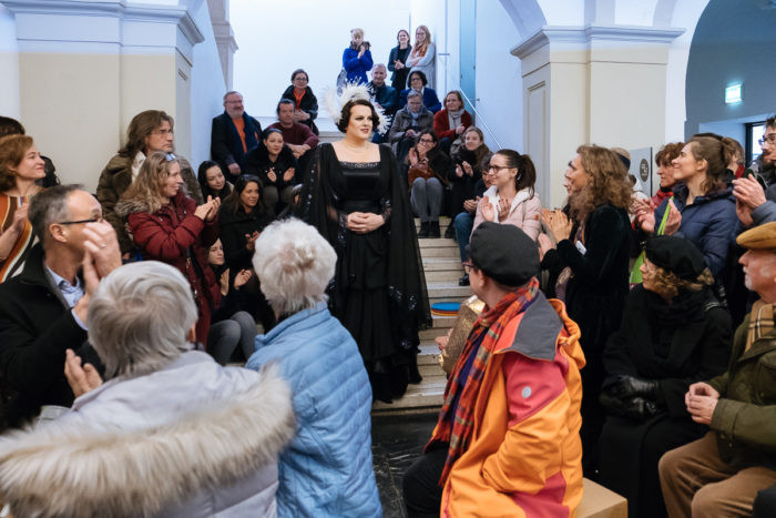 Eine Frau in einem schwarzen Kleid geht ganz normal anders durch eine sitzende, applaudierende Menge in einem Innenraum.