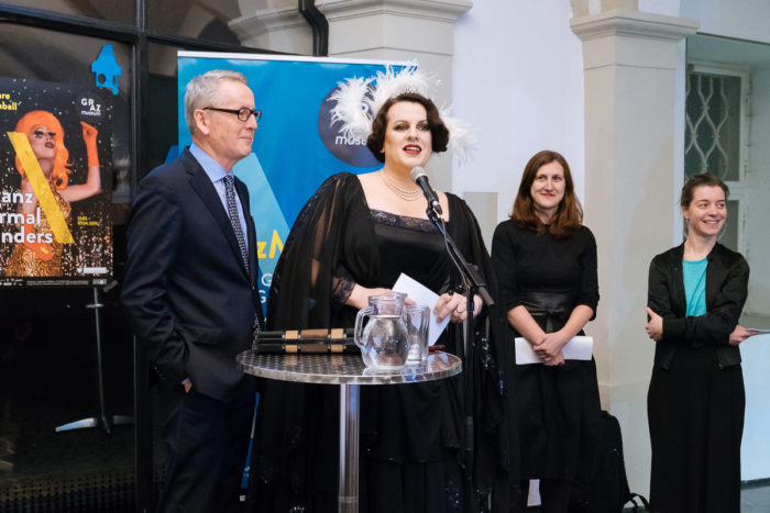 Vier Menschen, ganz normal anders, stehen an einem Podium, einer spricht in ein Mikrofon.