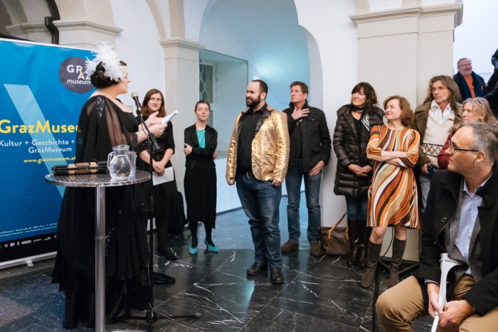 Eine kostümierte Person spricht bei einer ganz normal anders Indoor-Veranstaltung vor einem Grazer Museumsbanner.